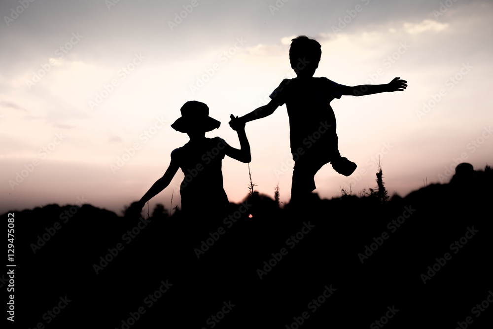 Silhouettes of kids jumping off a cliff at sunset. Boy and girl jump high holding hands. Brother and sister having fun in summer. Friendship, freedom concept. Twins on vacation in mountains.