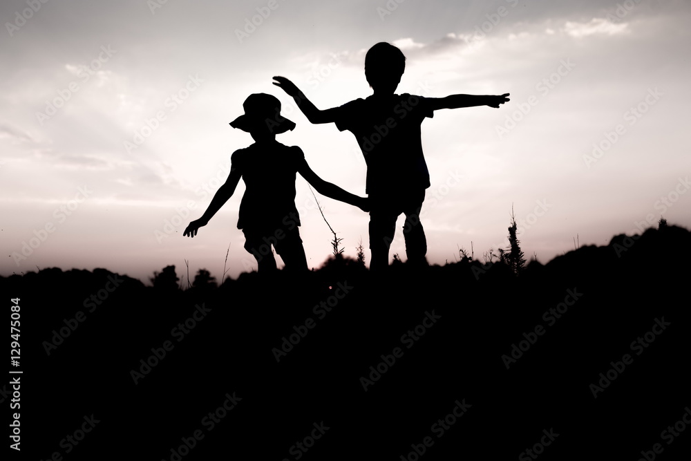 Silhouettes of kids jumping off a cliff at sunset. Little boy and girl jump raising hands up high. Brother and sister having fun in summer. Friendship, freedom concept. Twins on vacation
