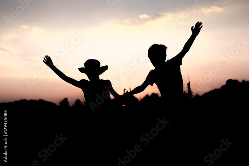 Silhouettes of kids jumping off a cliff at sunset. Boy and girl jump high holding hands. Brother and sister having fun in summer. Friendship, freedom concept. Vacation in mountains.