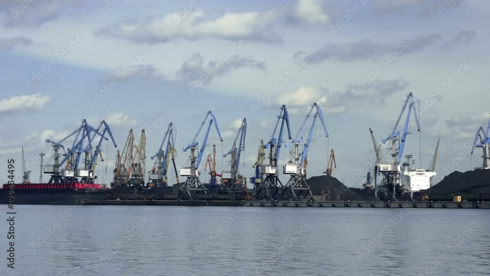 In the Port many cranes working on coal heaps, ship and clouds background