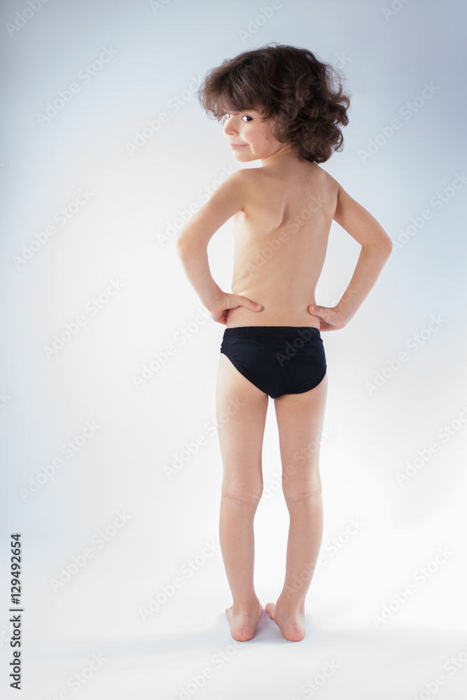 Curly boy in swimming trunks standing in full length with his back to the camera, looking at the camera and smiling. Gray background. Curly boy in swimming trunks standing in full length with his back to the camera, looking at the camera and smiling. Gray background.