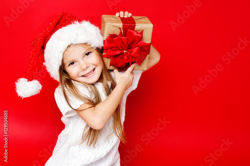 Happy smiling girl in Christmas cap with gift wrapped in craft paper and red ribbons on red background. Christmas. Happy family.