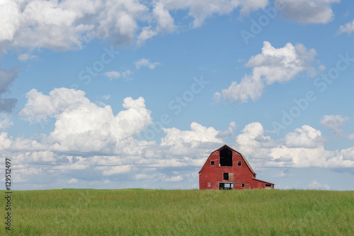 old abandoned red barn sitting in a field of green grass under a blue sky filled with white clouds