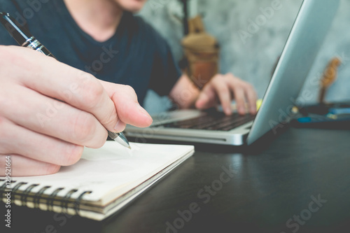 Casual young man writing some data in paper and using laptop on the desk