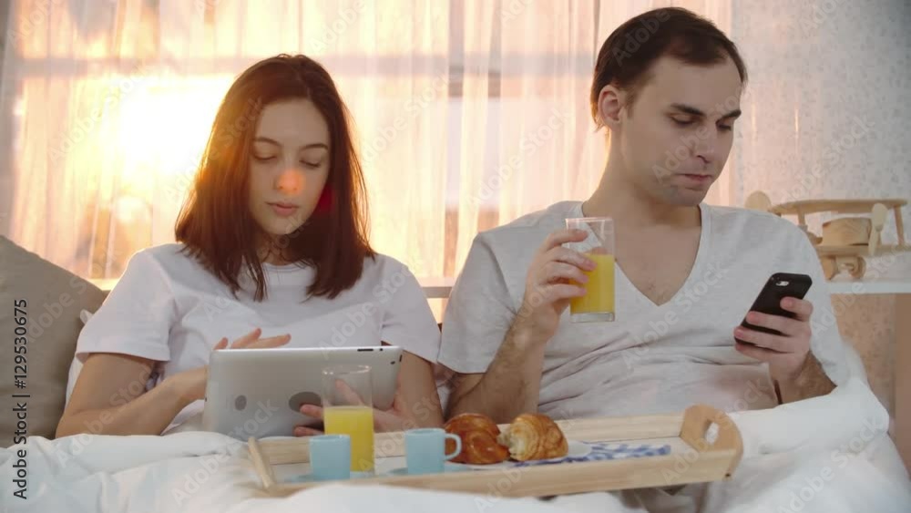 Young woman sitting in bed looking at tablet and drinking juice from breakfast tray alongside her husband with mobile phone Young woman sitting in bed looking at tablet and drinking juice from breakfast tray alongside her husband with mobile phone