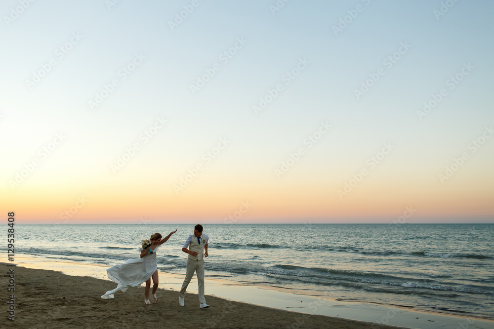 Laughing bride and groom run along the shore in evening lights