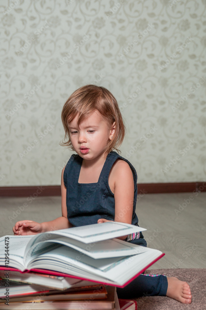 A little girl in a dark blue dress reading a book sitting on the floor near teddy bear. Child reads story for toy. Turns the page. A little girl in a dark blue dress reading a book sitting on the floor near teddy bear. Child reads story for toy. Turns the page.