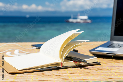 Opened empty notepad is on the table with phone and headphones at the tropical sea background