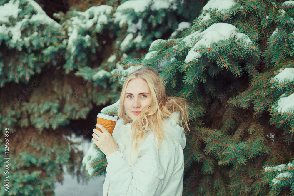 Young beautiful woman with blond hair wearing a white jacket, standing in a cold winter day holding hot coffee or tea. Behind the woman background trees and snow. Young beautiful woman with blond hair wearing a white jacket, standing in a cold winter day holding hot coffee or tea. Behind the woman background trees and snow.