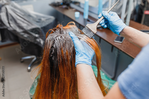 Canvastavla In the professional salon stylist in gloves with brush applying paint to the bright red hair of the client