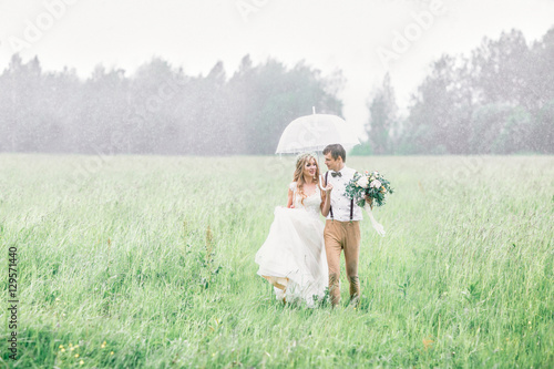 The blonde bride and groom are on the field in the rain. Wedding on a rainy day.