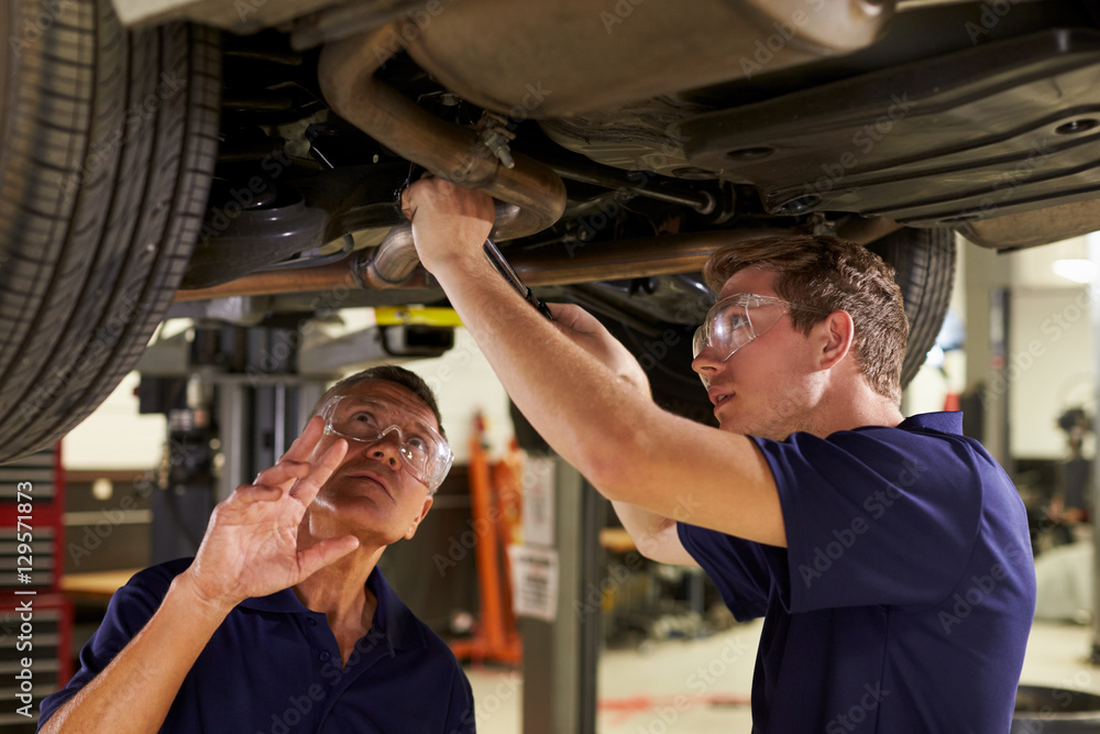 Mechanic And Male Trainee Working Underneath Car Together Stock Photo ...