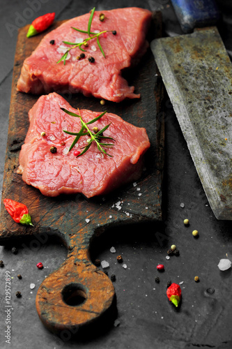 raw beef steak on rustic cutting board, and rustic old knife for meat, rosemary salt and pepepr chili on a dark background.selective focus photo