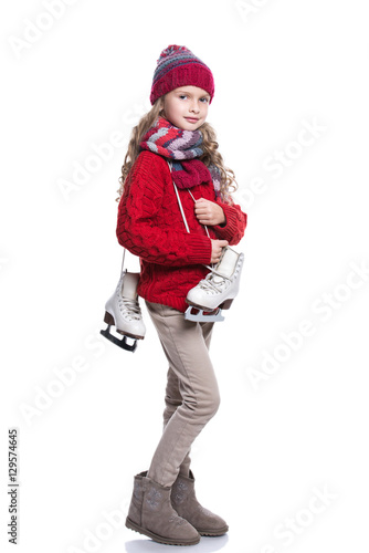 Cute smiling little girl with curly hairstyle wearing knitted sweater, scarf, hat and gloves with skates isolated on white background. Winter clothes and sport concept.