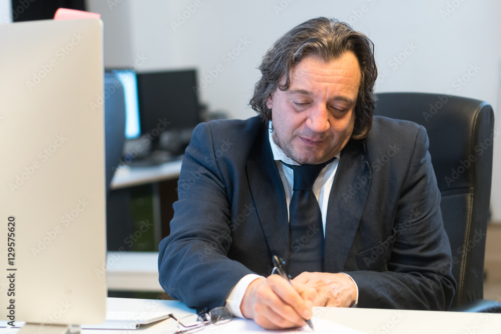 Businessman in suit and tie filling in some paper documents sitting at his working place Businessman in suit and tie filling in some paper documents sitting at his working place