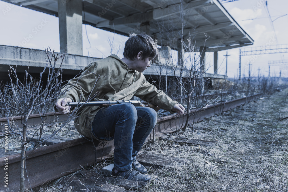 young boy sitting on the railroad tracks near the abandoned train station. wandering boy with a gun. man in a protective cloak with a hood. Post apocalypse. traveling on foot in a post-apocalyptic young boy sitting on the railroad tracks near the abandoned train station. wandering boy with a gun. man in a protective cloak with a hood. Post apocalypse. traveling on foot in a post-apocalyptic