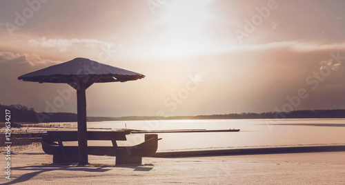 Wooden bench with umbrella on lake cowered with snow at beautiful sunset in winter evening. Focus point on the bench and umbrella. © fotoduets