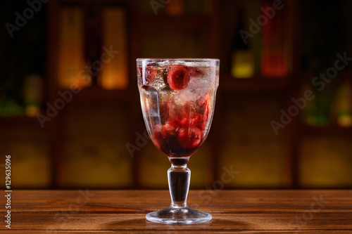 Cocktail with alcohol, cherries and ice cubes on a wooden table with blurred background of bottles and light. 