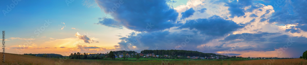 Sunset in the countryside. Dramatic sky with clouds, field and rural houses on a background of trees. Panoramic shot. Sunset in the countryside. Dramatic sky with clouds, field and rural houses on a background of trees. Panoramic shot.