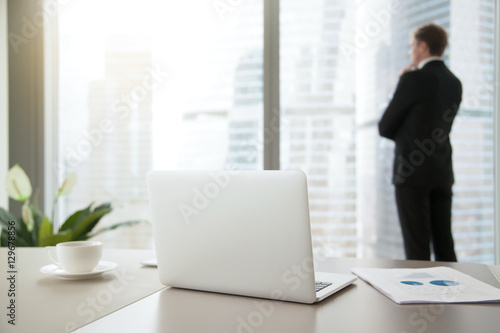Company president in formal suit standing, looking through window at cityscape, dreaming, planning new projects, resting after intensive work. Focus on laptop on office table. Business success concept