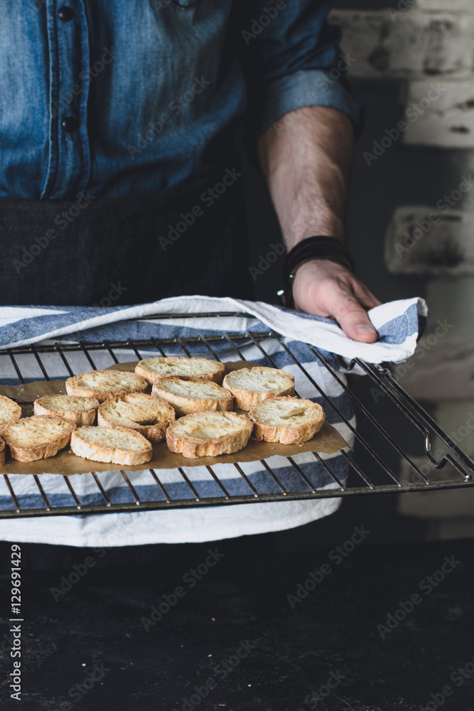 Man holding cooling rack with fresh from the oven crostini, sliced and baked italian baguette with olive oil. Vertical composition, selective focus, toned image.