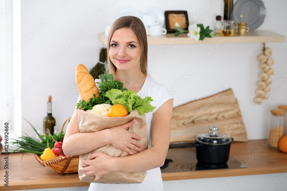 Young cheerful smiling woman is ready for cooking in a kitchen. Housewife is holding big paper bag full of fresh vegetables and fruits and  looking at the camera