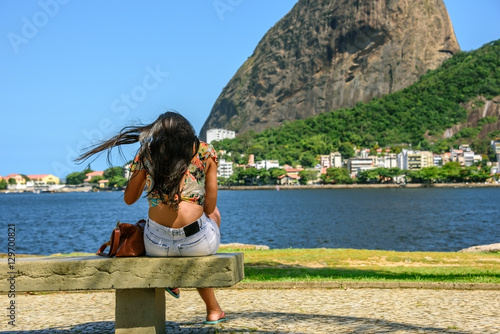 Beautiful woman with flying black hair and brown bag sitting on the bench on the background of Sugarloaf and Guanabara bay in Rio de Janeiro, Brazil