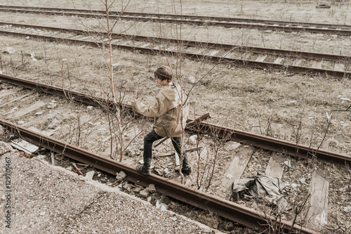 young boy walking on railroad tracks. He is holding the gun. wandering boy. man in a protective cloak with a hood. Post apocalypse. traveling on foot in a post-apocalyptic world.