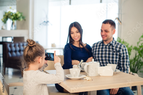 family, parenthood, technology people concept - close up of happy mother, father and little girl having dinner, kid taking photo by smartphone at restaurant