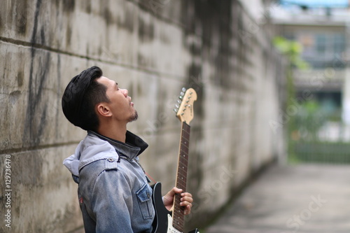 A young man wearing jeans, playing a guitar on a brick wall back