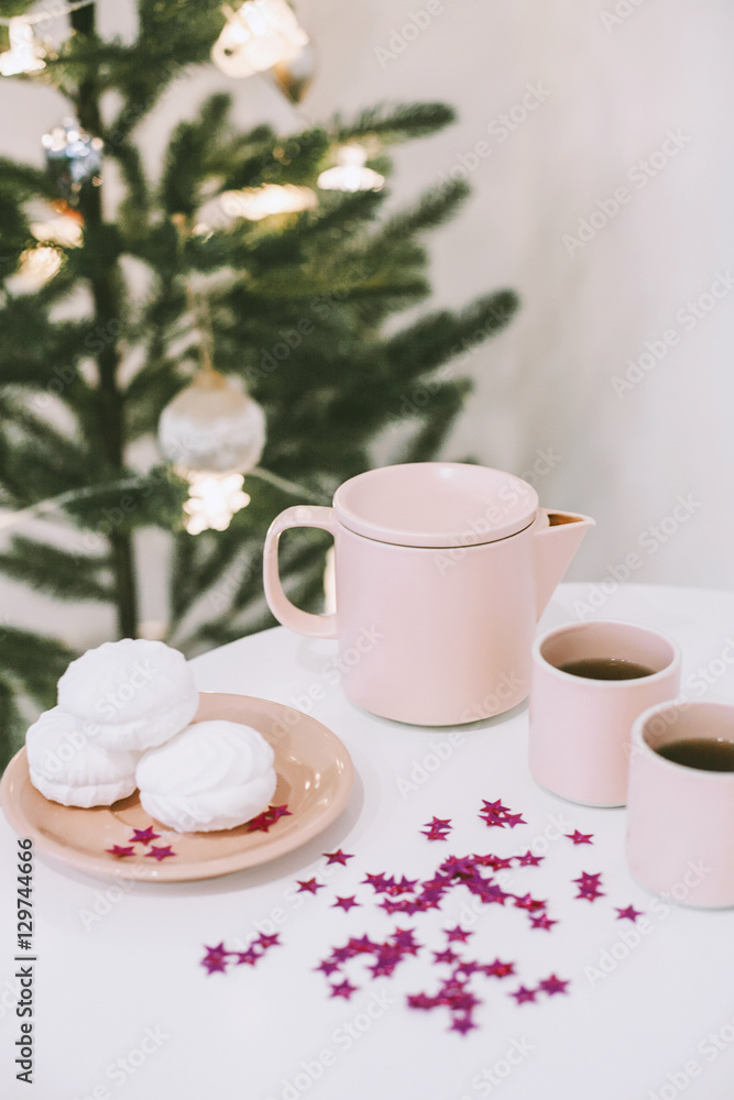 White table with a pink teapot and mugs and some tasty marshmallows on a plate in a white room near the christmas tree