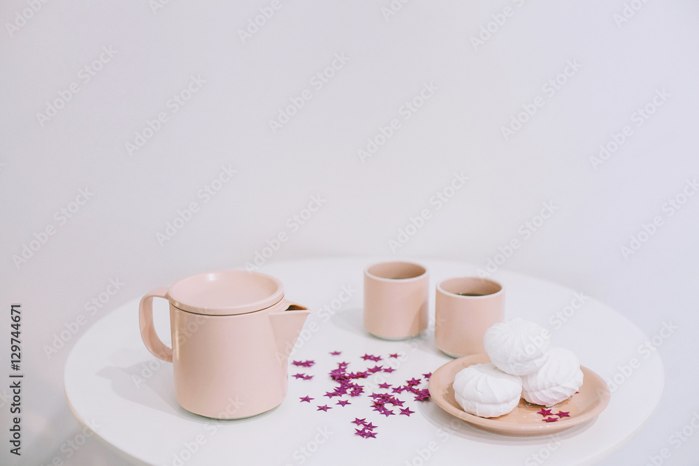 White table with a pink teapot and mugs and some tasty marshmallows on a plate in a white room White table with a pink teapot and mugs and some tasty marshmallows on a plate in a white room