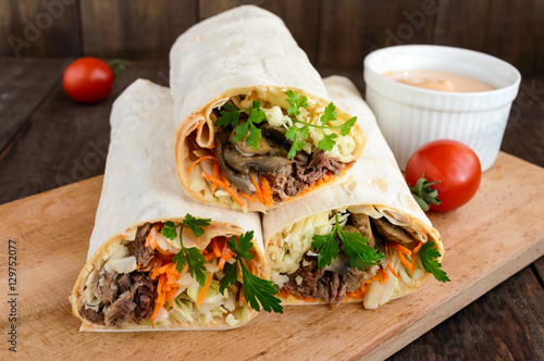 Shawarma sandwich - fresh roll of thin lavash (pita bread) filled with grilled meat, mushrooms, cheese, cabbage, carrots, sauce, green. Traditional Eastern snack. On a dark wooden background.