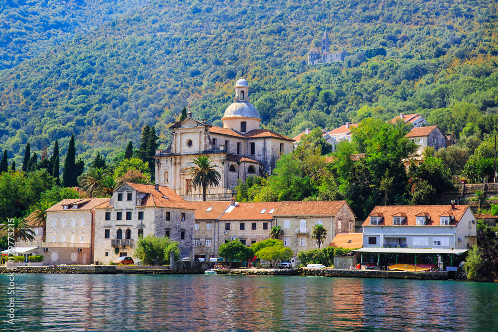 Waterfront of small town Prcanj along Bay of Kotor, Montenegro. View of Birth of Our Lady Church, coastal villas, gardens and mountain from sea.