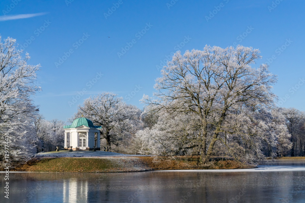 Idyllische winterliche Parklandschaft in Kassel, Karlsaue, Deutschland, auf einer kleinen Insel im See liegt ein kleiner Tempel, der See ist halb zu gefroren und leichter Schnee bedeckt die Bäume