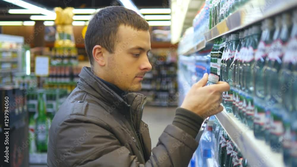 Young man choosing and buying bottle of mineral water at the supermarket. Guy taking product from shelves at grocery section of shop. Man buys drinks in store. Close up Young man choosing and buying bottle of mineral water at the supermarket. Guy taking product from shelves at grocery section of shop. Man buys drinks in store. Close up