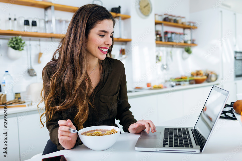 Beautiful young woman working with laptop while eating in the kitchen.