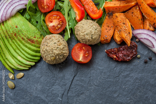 Vegan food frame: avocado, sweet potato, lentil cutlets, tomatoes, arugula, onions on black stone background with a copy space in the center
