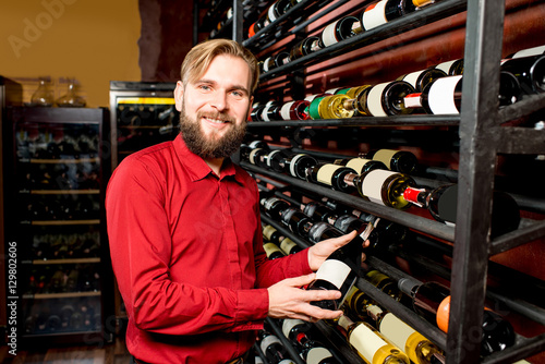 Portrait of a handsome sommelier with glass of wine in front of the shelves with bottles at the luxury supermarket or restaurant