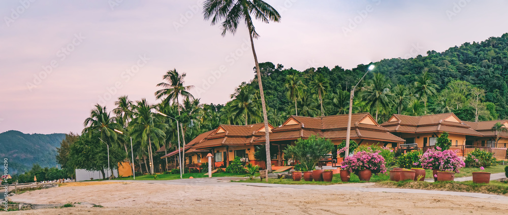 Panoramic view of tropical hotel beach among the palm trees, beautiful lawn and benches at sunset. Beach sunset is a golden sunset sky with a wave rolling to shore