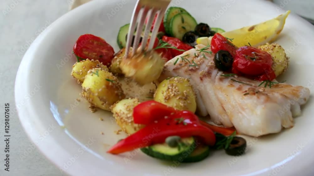 A meal of fish on a plate with potatoes and tomatoes. Fork picking up a potatoe. A meal of fish on a plate with potatoes and tomatoes. Fork picking up a potatoe.