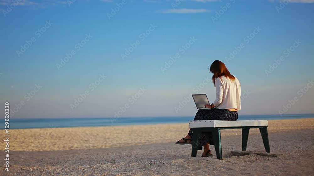 caucasian girl writes a text on a laptop sitting on a bench on the beach, bubbles are flying along the seashore hd caucasian girl writes a text on a laptop sitting on a bench on the beach, bubbles are flying along the seashore hd