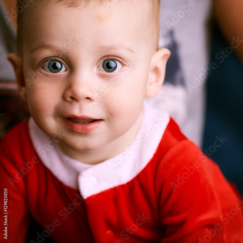 Square image of baby boy dressed in a Santa Claus outfit smiling at the camera with beautiful blue eyes