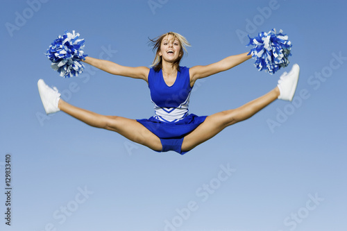 Full length of excited cheerleader with pompoms doing splits against blue sky photo
