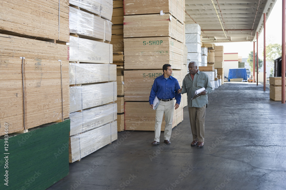 Two multiethnic men with clipboards walking along stacks of wood in warehouse Two multiethnic men with clipboards walking along stacks of wood in warehouse
