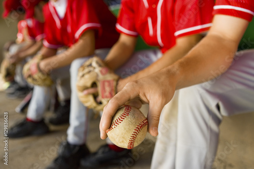 Low section of baseball team mates sitting in dugout with player holding a ball in foreground