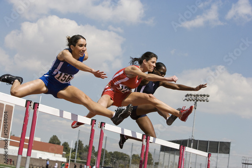 Multiracial female athletes clearing hurdles in race photo