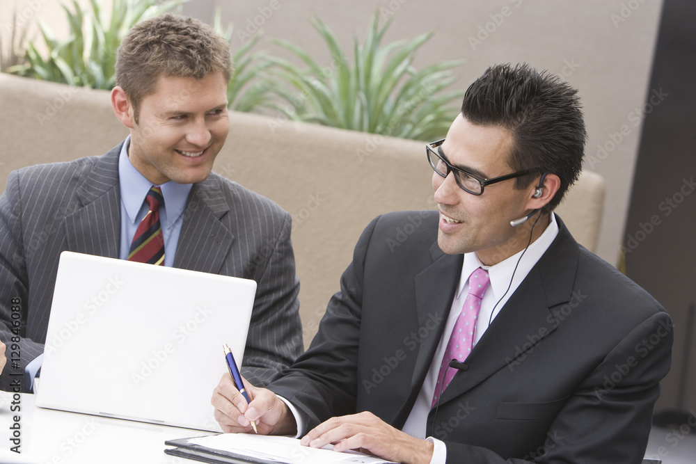 Happy businessman using laptop with colleague writing on document Happy businessman using laptop with colleague writing on document