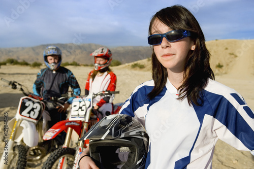 Young female off road motor biker holding helmet with friends in the background at track