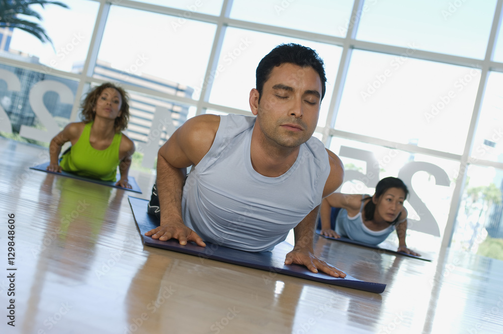 Man with eyes closed exercising on mat with women in the background Man with eyes closed exercising on mat with women in the background